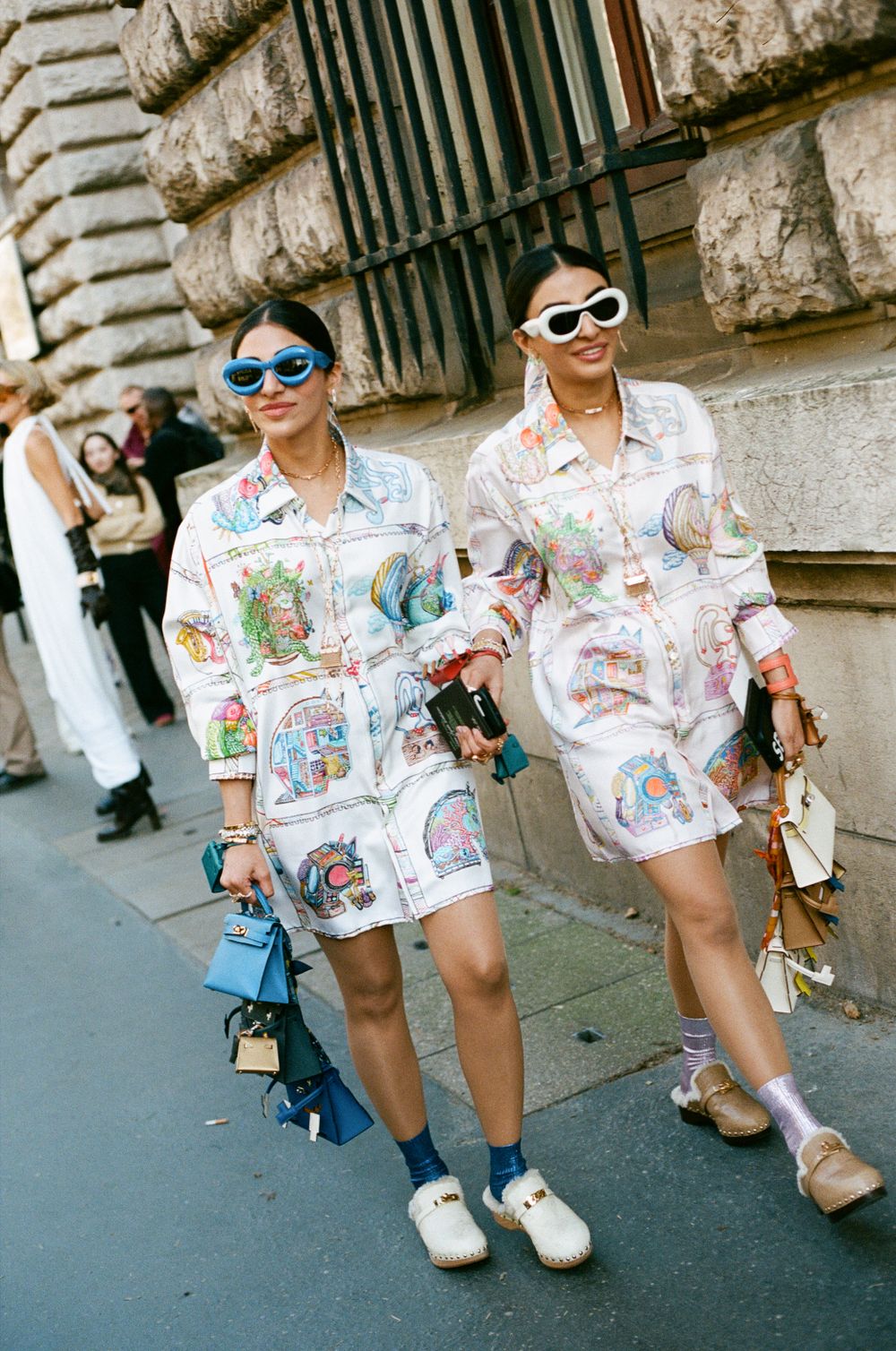 Two fashionable people wearing matching printed oversized shirt dresses and sunglasses walk together on a city street.