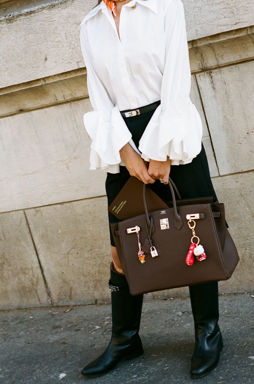 Stylish outfit featuring white ruffled blouse, black pants, boots and brown leather Hermes Birkin bag with decorative charms.