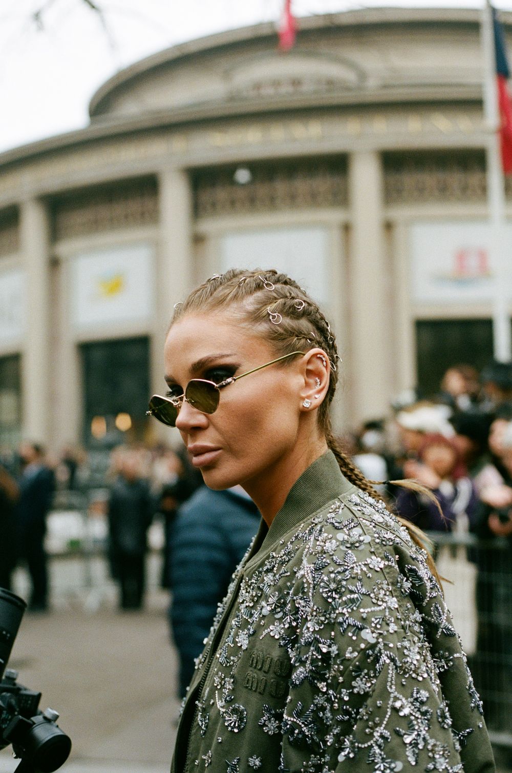 Fashion attendee wearing sunglasses and braided hair with an embellished olive bomber jacket outside an event venue.