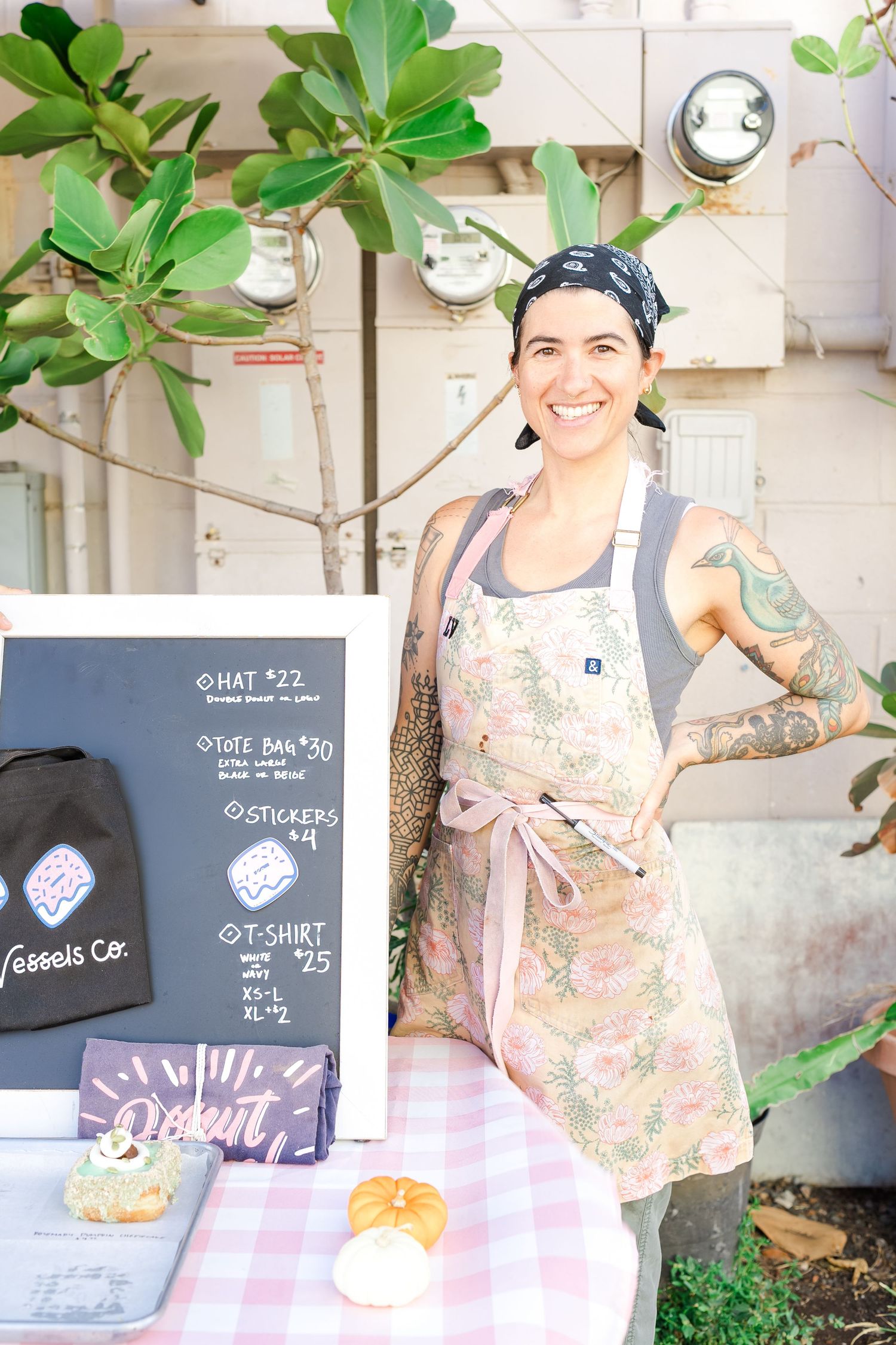 A person in a floral apron stands at a food stall with a menu board and small desserts displayed on a checkered tablecloth.