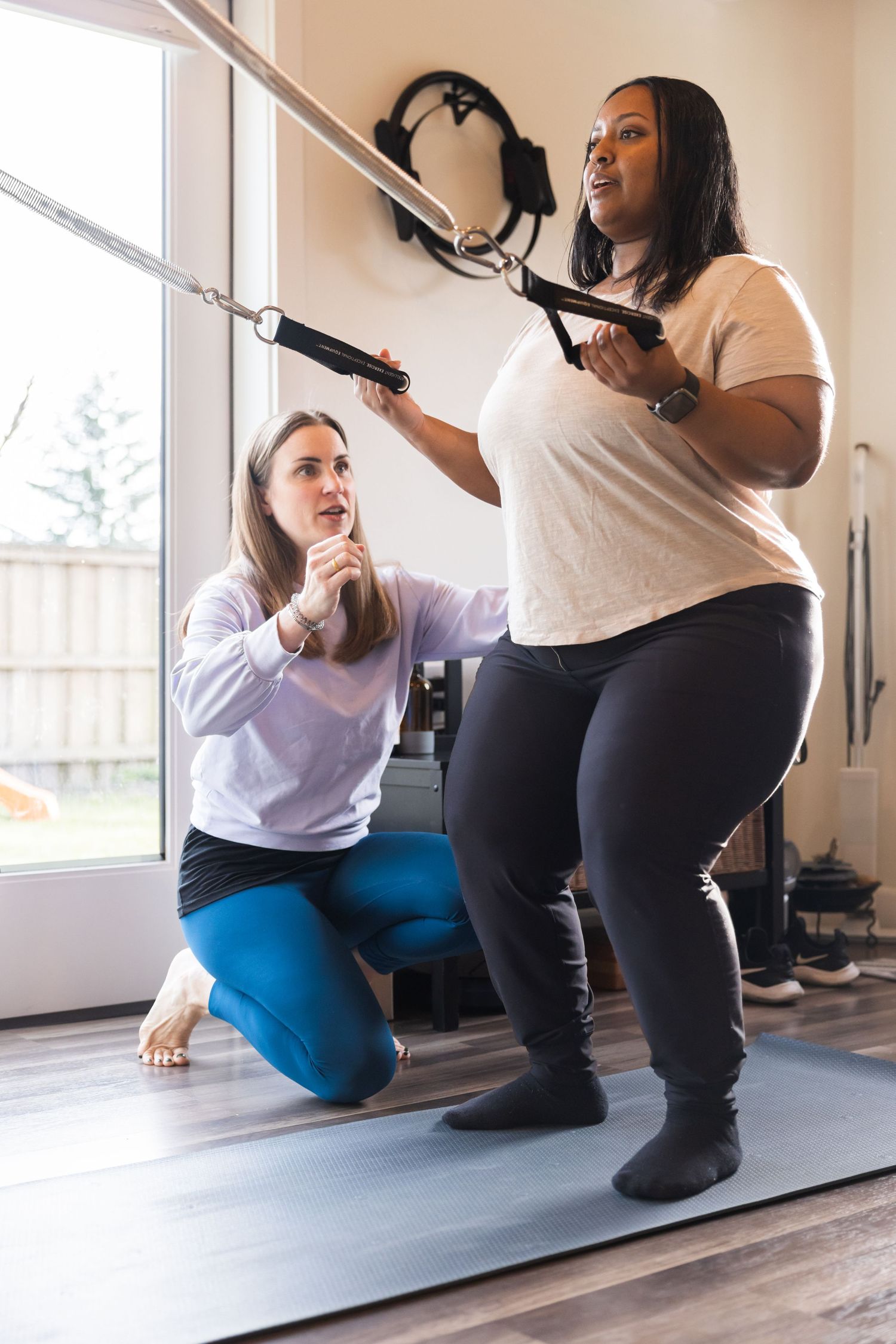 Two people exercising together with resistance bands in a home gym space with natural lighting through a window.