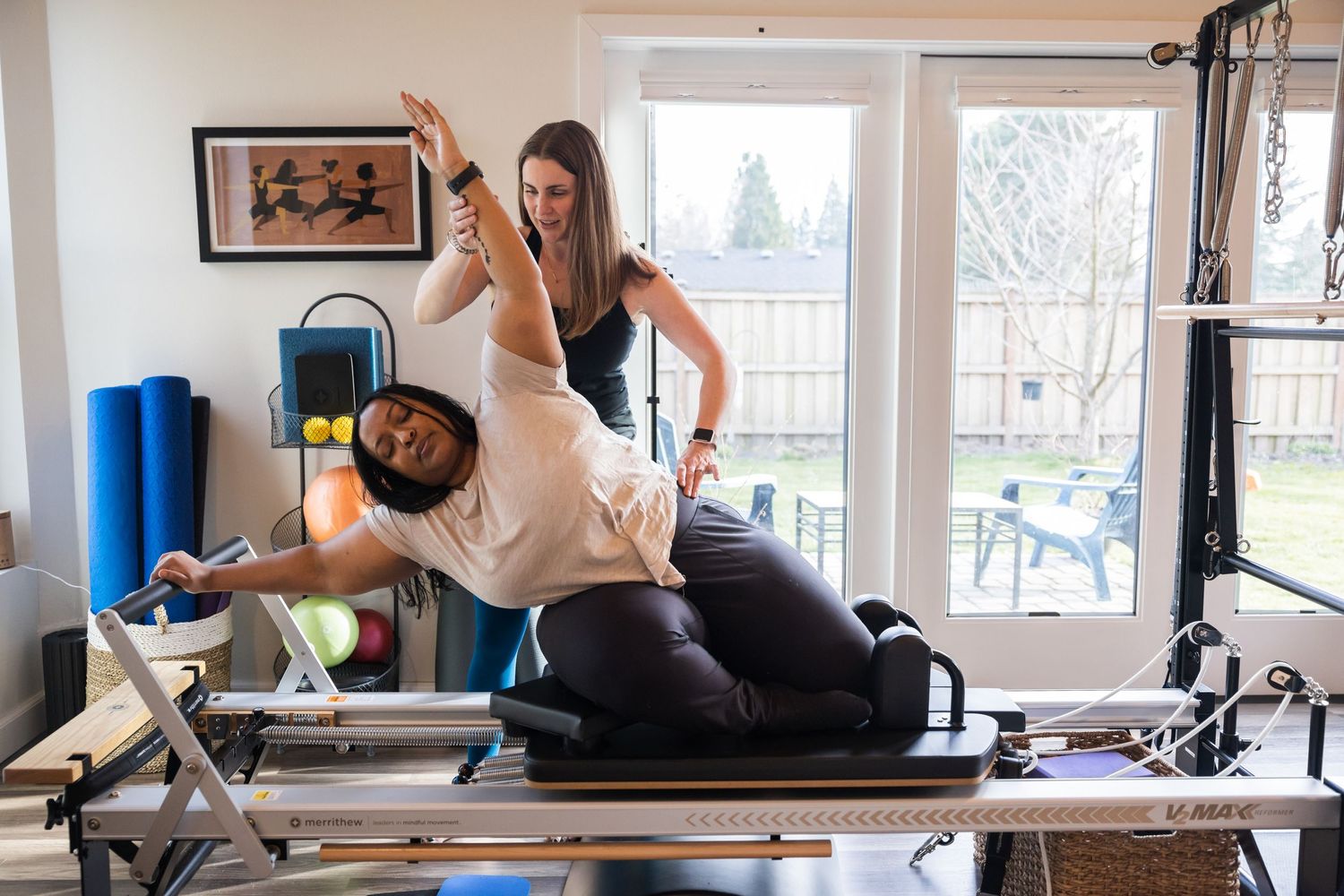 Pilates instructor guides client through stretching exercise on reformer machine in sunlit home studio.