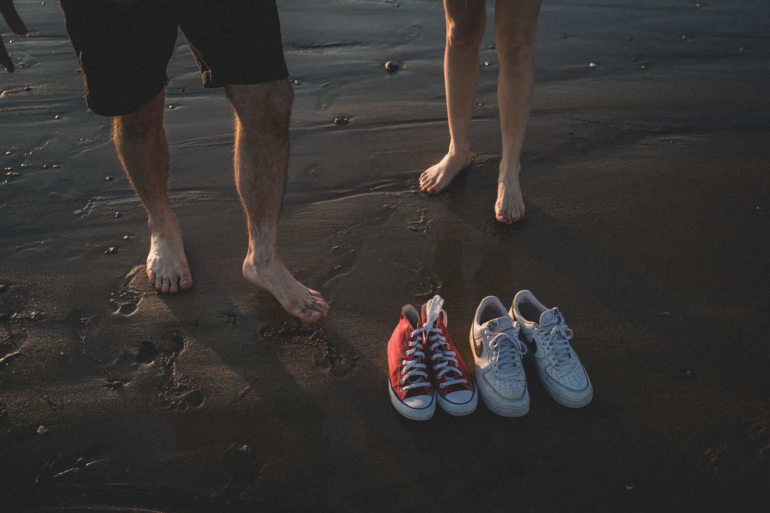 Bare feet standing on a beach next to colorful sneakers on dark sand.
