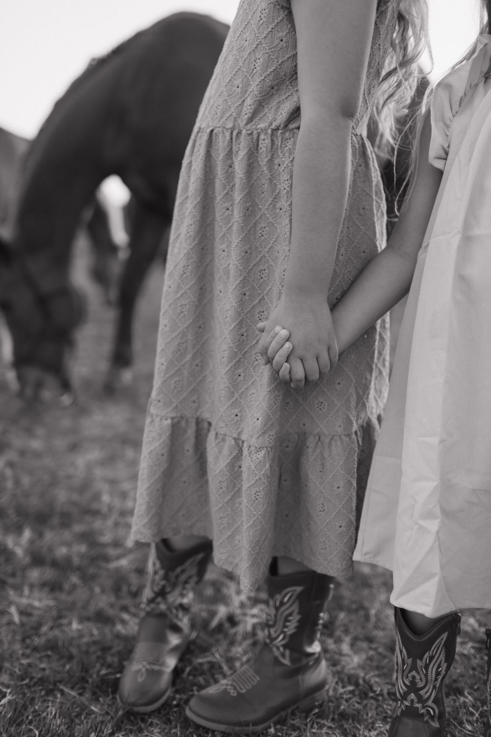 Black and white detail shot of western boots and flowing dresses with horses grazing in background.