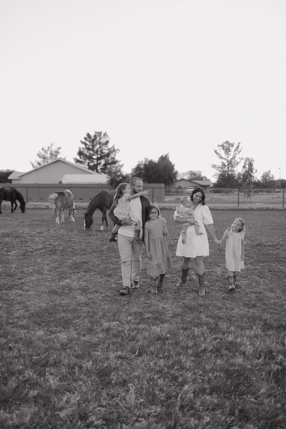 Black and white portrait of a family standing in a field with horses behind them at dusk.