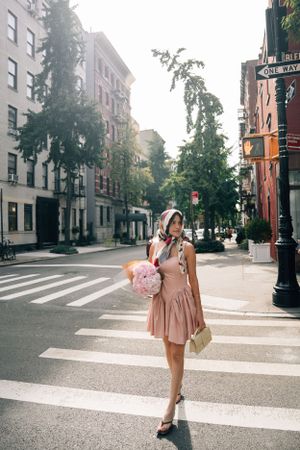 Person in pink dress holding balloons crosses a tree-lined street in urban neighborhood on sunny day.
