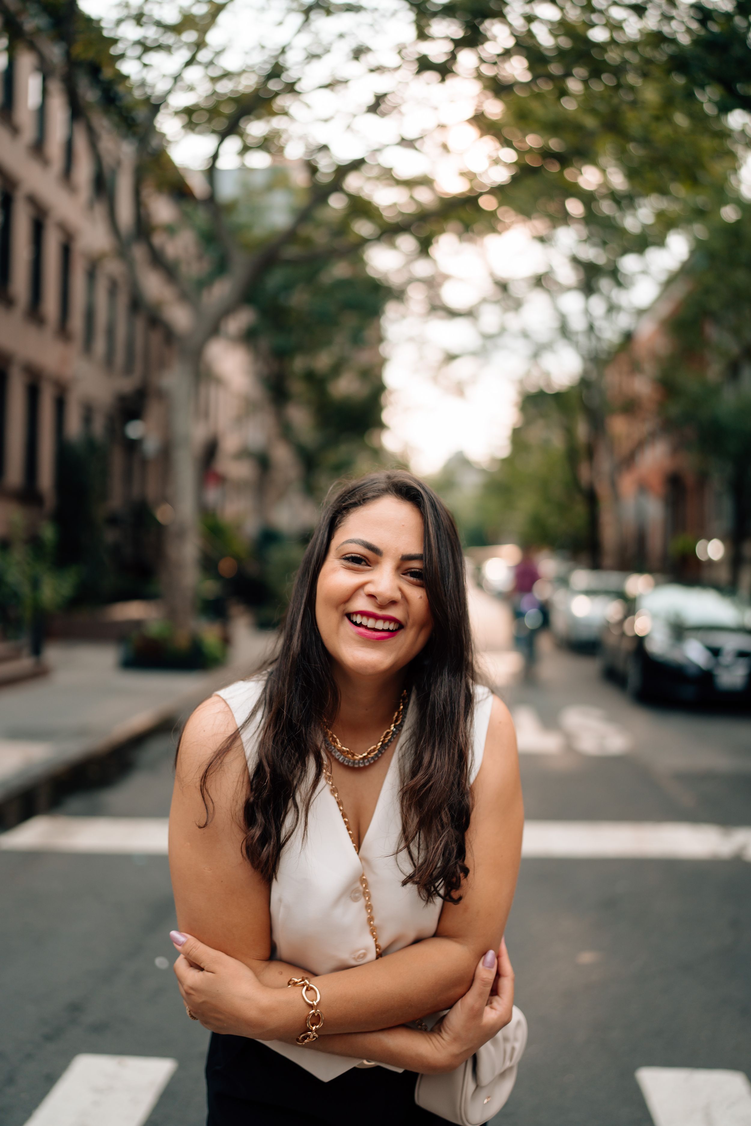 Person in a white sleeveless top laughs warmly while standing on a tree-lined city street at sunset.