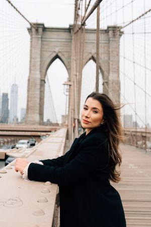 Someone in a black coat poses against the iconic stone arches and cables of the Brooklyn Bridge in New York City.