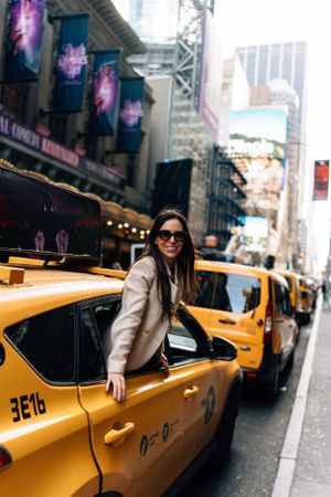 A woman in a beige coat and sunglasses getting into a yellow NYC taxi on a busy city street.