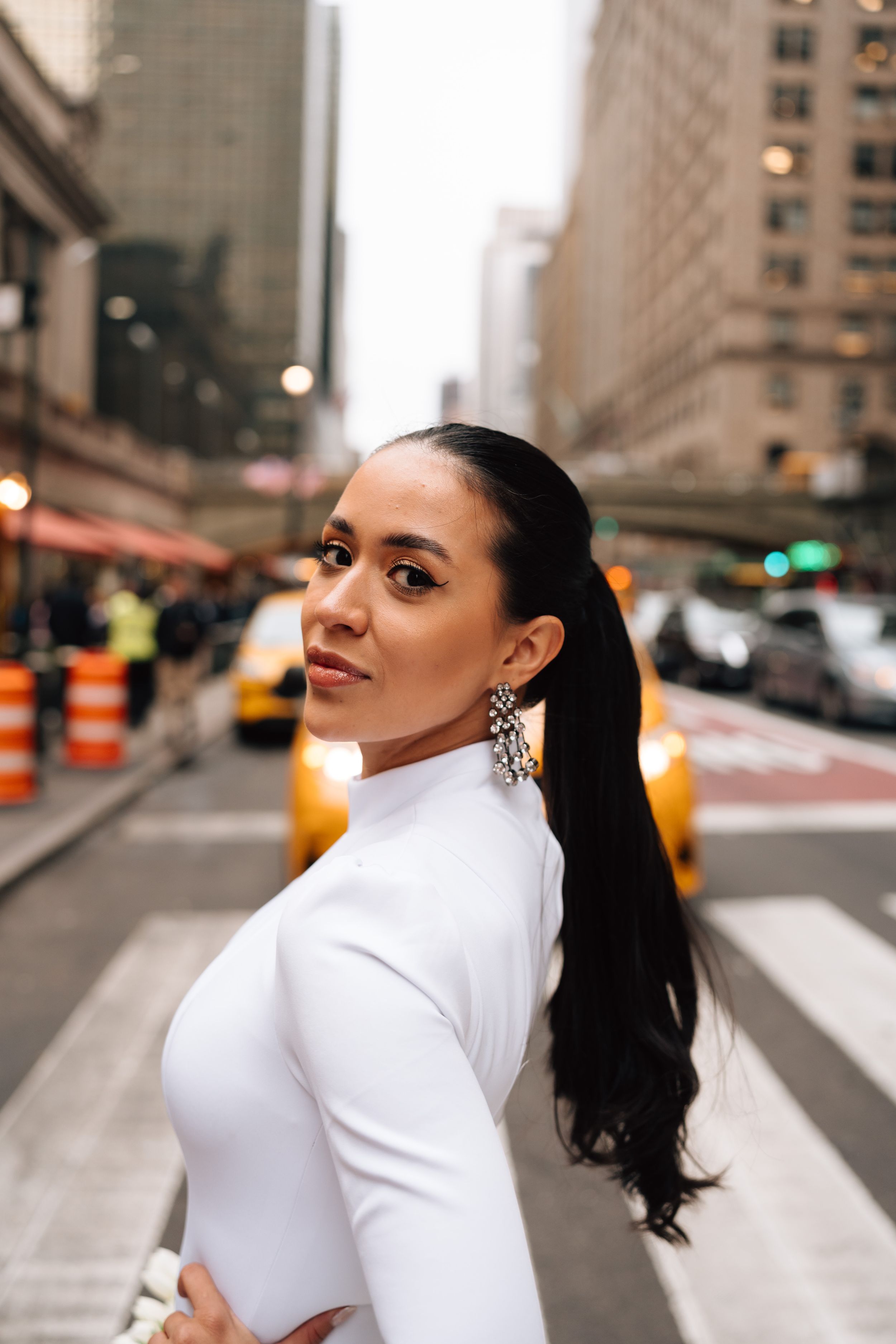 A person in a white turtleneck poses on a city street with yellow taxis and tall buildings in the background.