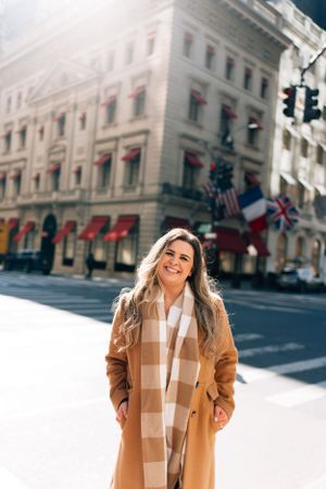 Woman wearing a camel coat and plaid scarf stands on a city street with international flags on a building behind her.