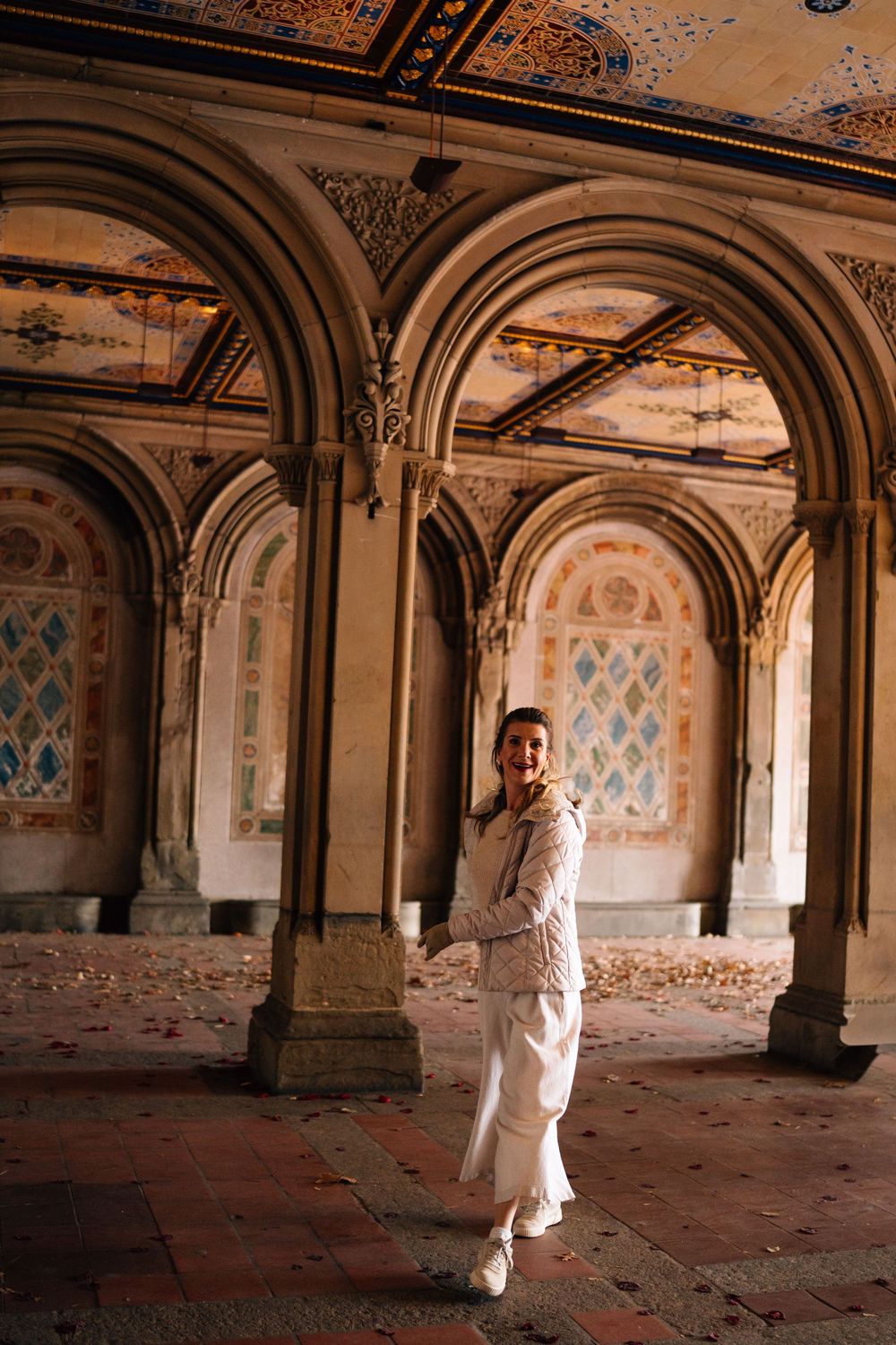 Figure in white dress stands in ornate abandoned Bethesda Terrace arcade with arched ceilings and peeling paint.