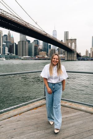 A person in jeans and white top poses on a wooden deck with Brooklyn Bridge and Manhattan skyline in the background.
