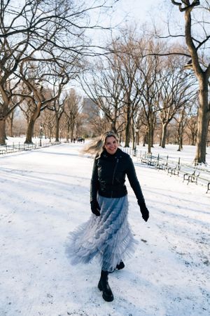 A person wearing a flowing skirt and winter attire twirls in the snow at Central Park surrounded by bare trees.