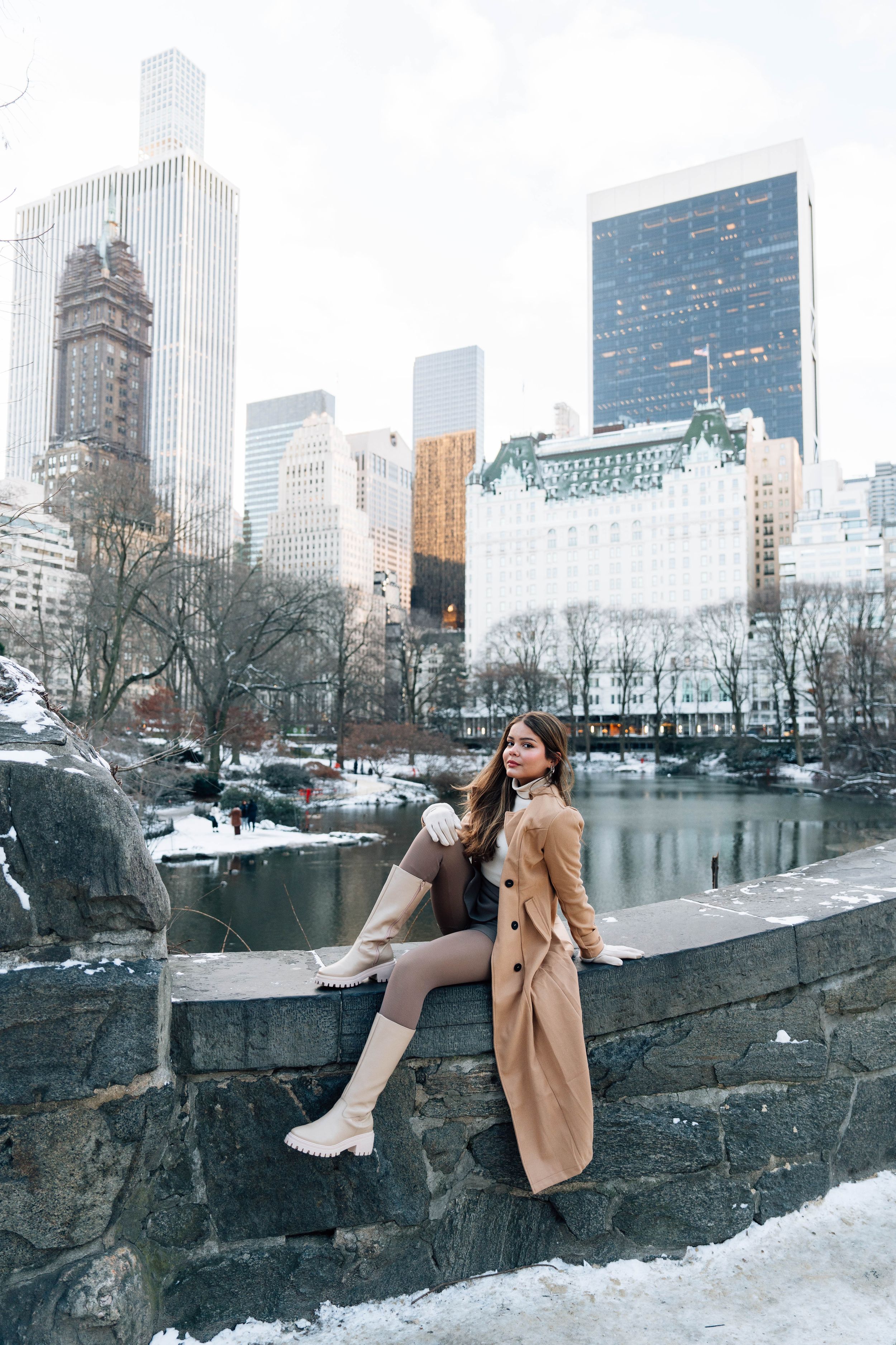 Winter scene at Central Park in New York City with snowy rocks, frozen pond and iconic buildings in background.