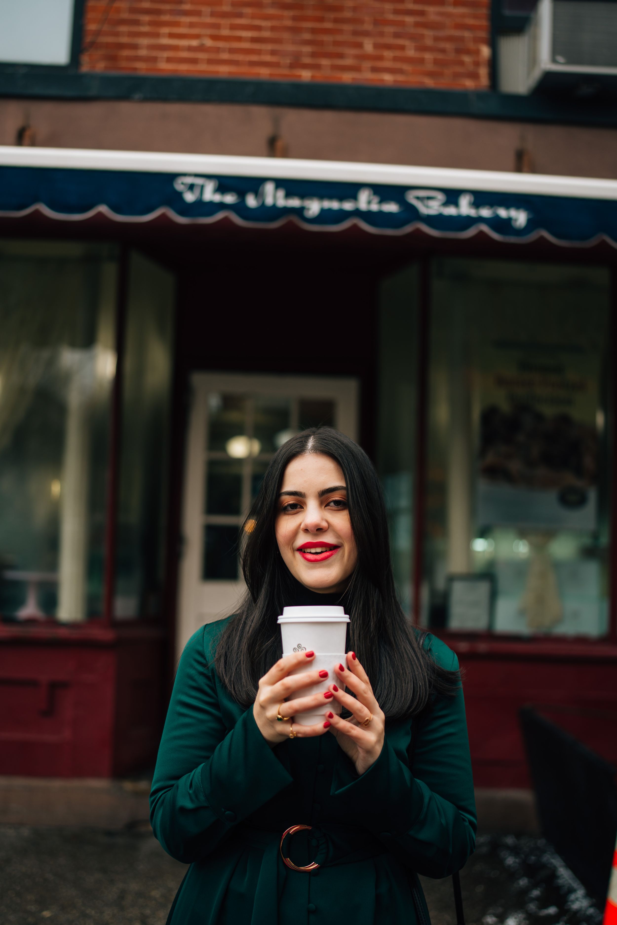 Someone wearing a green winter coat holds a coffee cup while standing in front of The Magnolia Bakery storefront.