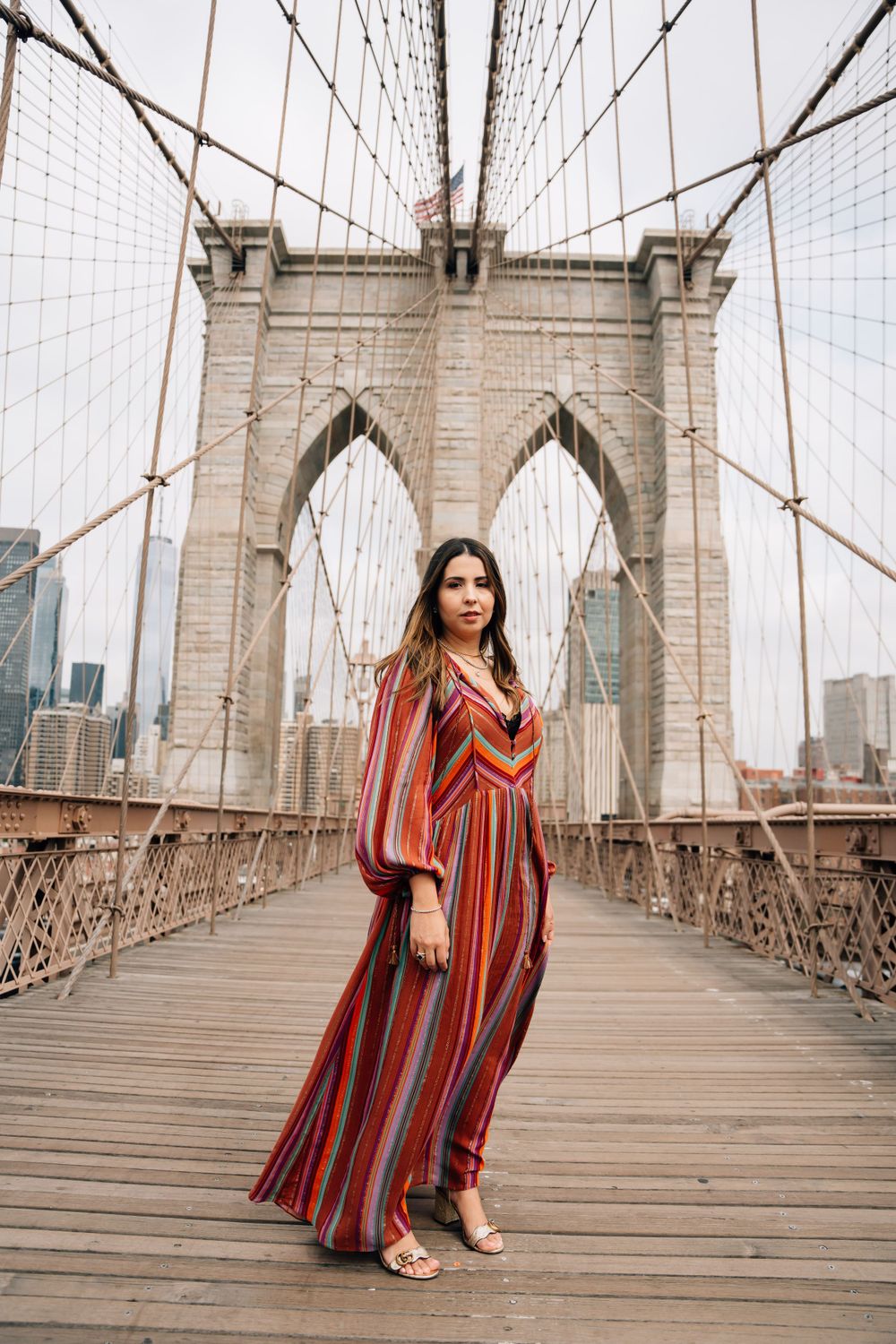 Woman in striped maxi dress poses on Brooklyn Bridge with Manhattan skyline in background.