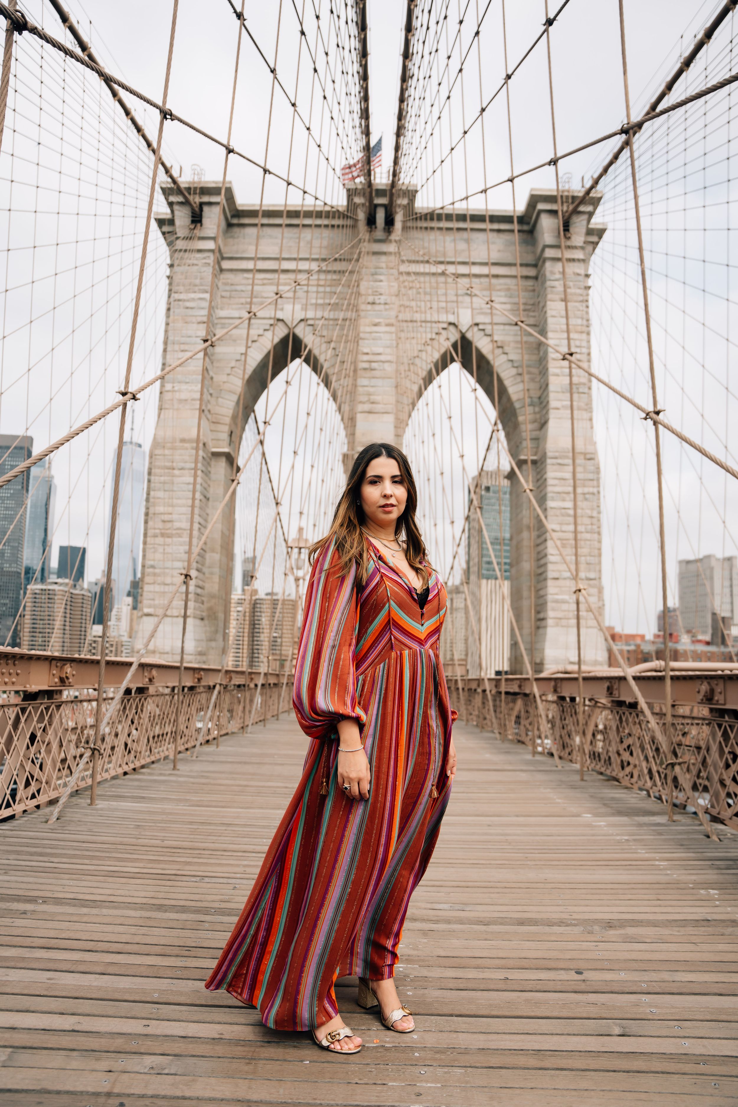 Woman in striped maxi dress poses on Brooklyn Bridge with Manhattan skyline in background.