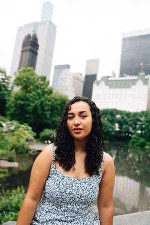 Person with curly hair in floral dress poses at Central Park with Manhattan skyline and skyscrapers in background.