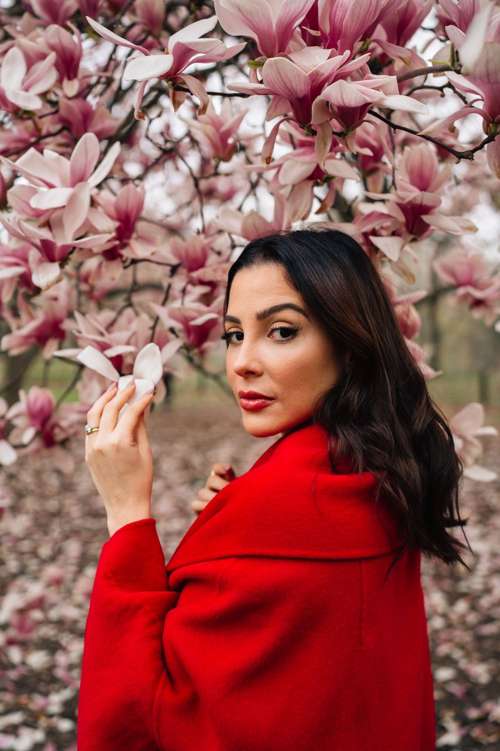 A person in a vibrant red coat poses elegantly against blooming pink magnolia flowers in spring.