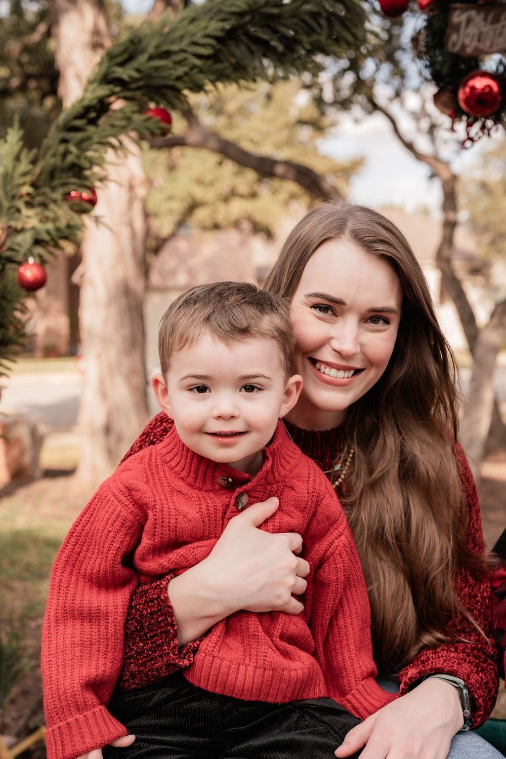 A mother and young child wearing matching red sweaters share warm embraces and smiles during an outdoor photo session.