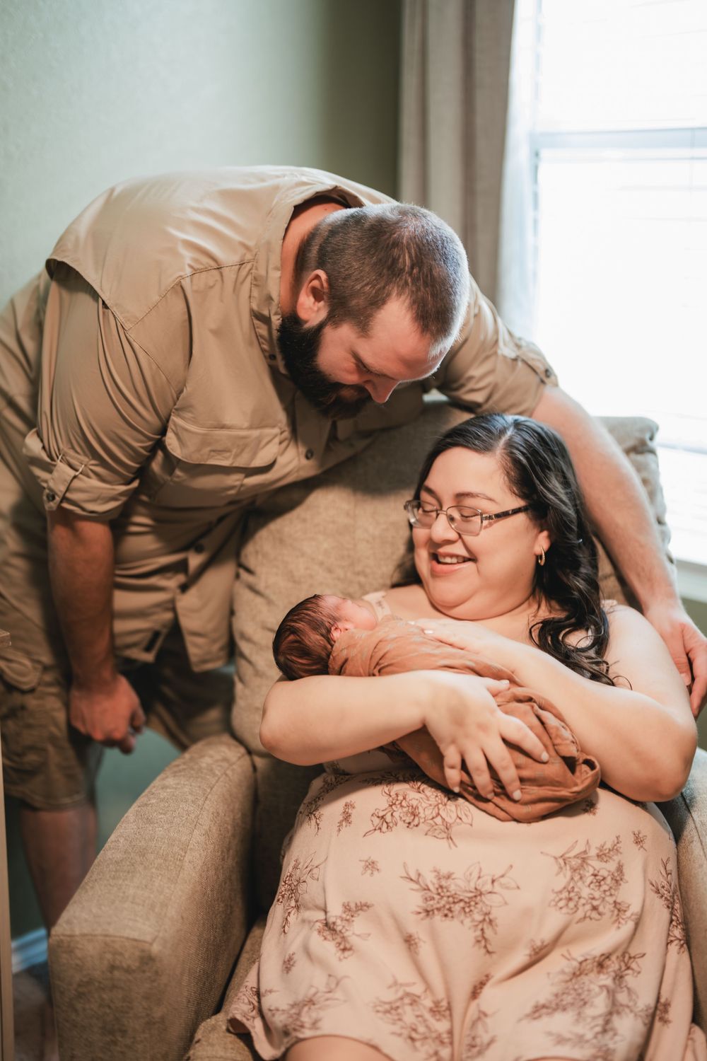 A tender family moment as parents lovingly gaze at their newborn baby while sitting on a beige couch near a window.