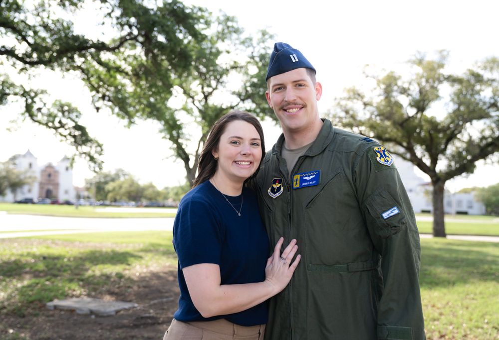 Military service member in uniform and companion pose together outdoors on a sunny day near oak trees.