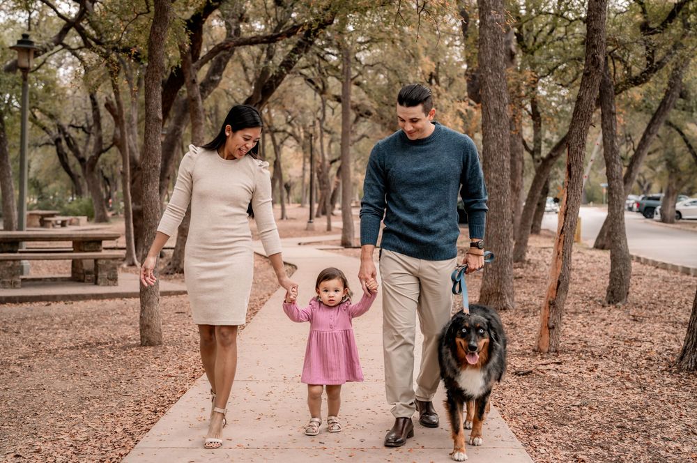 Family and dog walking together on a leaf-covered trail through a wooded park on an autumn day.