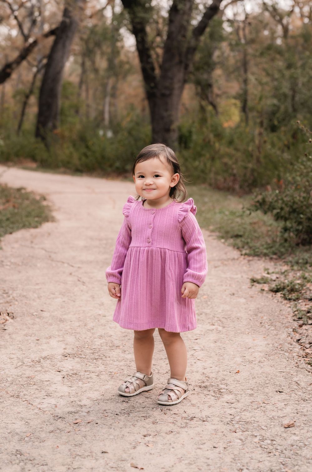 Small child in a pink ruffled dress standing on a dirt path surrounded by trees.