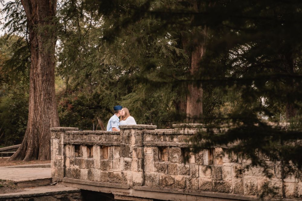 A couple poses for wedding photos in a rustic outdoor setting with stone walls and tall trees in the background.