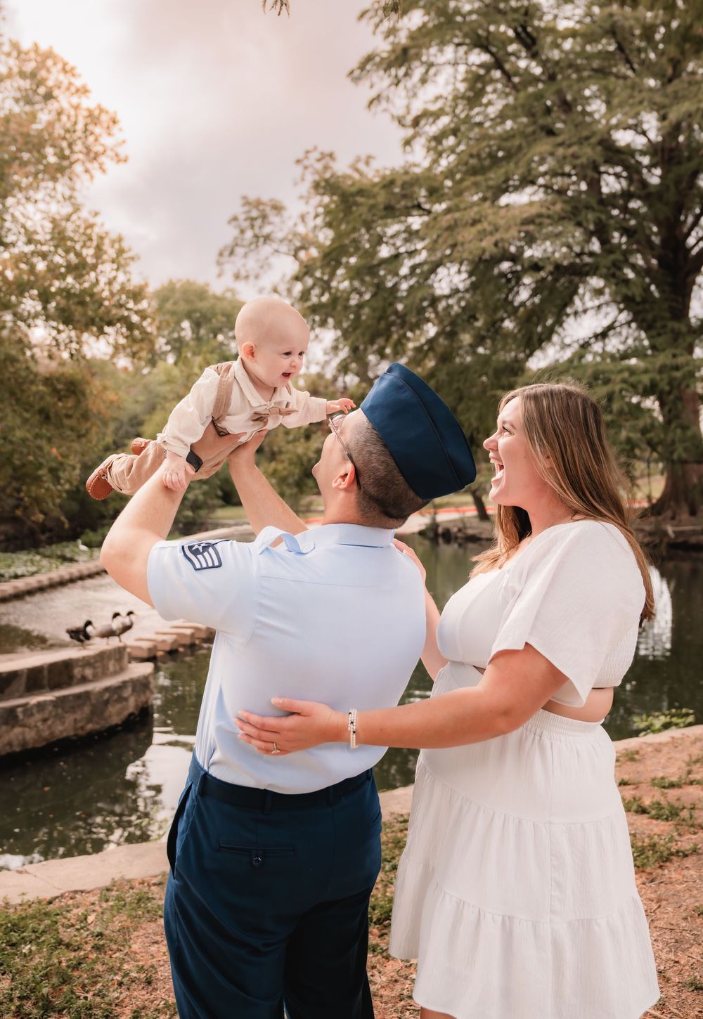A couple in white and navy outfits share a tender moment while holding a baby outdoors by a lake at sunset.