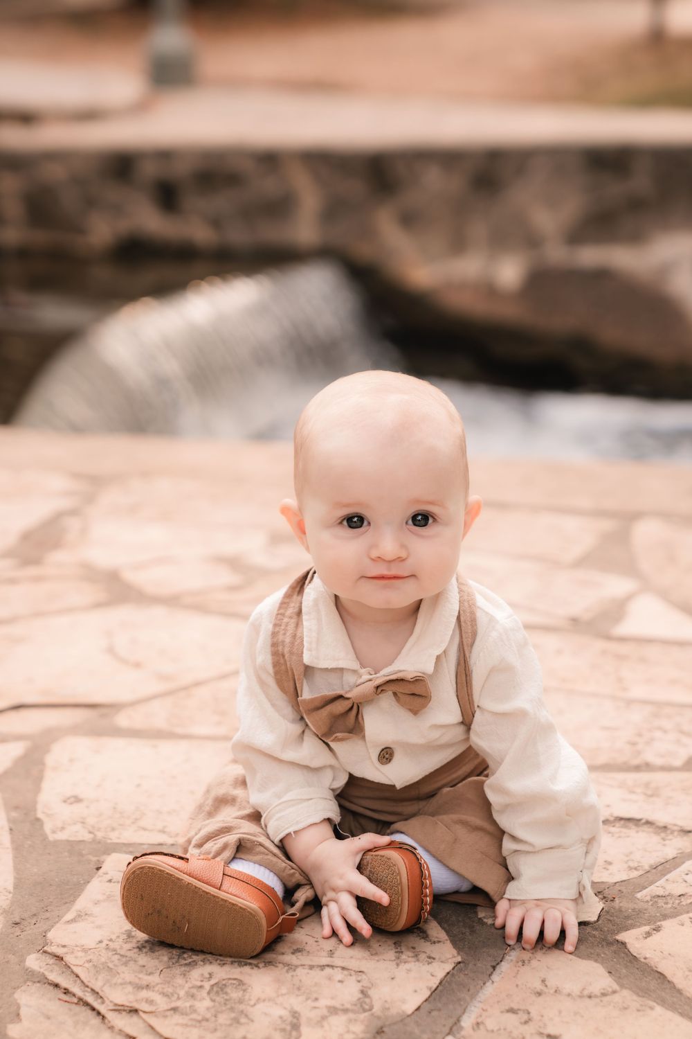 Baby in vintage-style outfit with white shirt, brown overalls and leather shoes sits on stone patio.