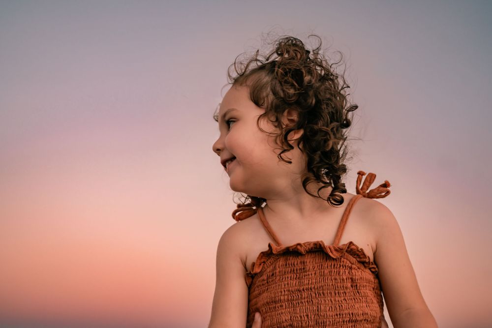 A young person with curly hair wearing a brown top smiles joyfully against a pink sunset sky.