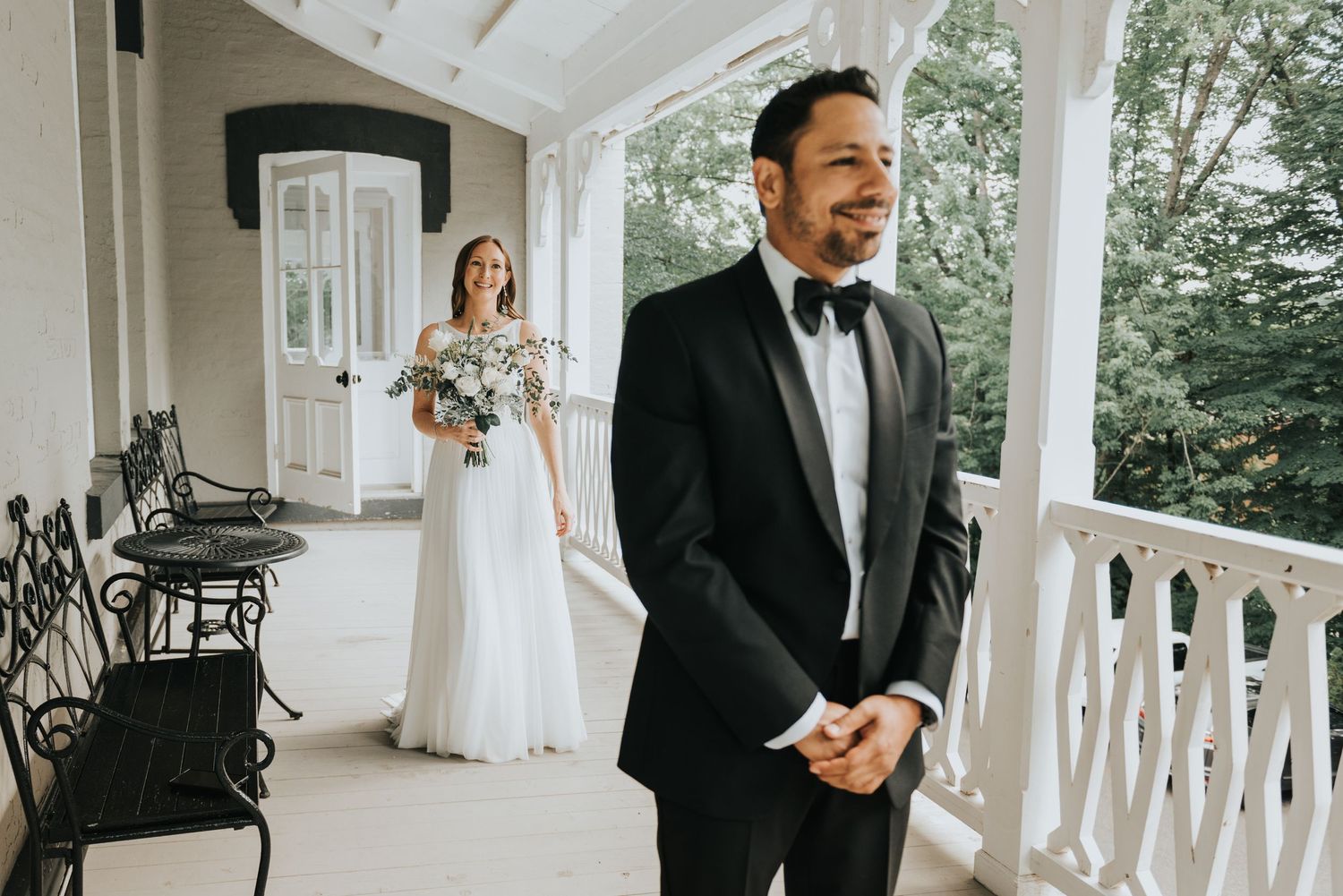 First look between bride in flowing white gown and groom in black tuxedo on elegant white porch during wedding day.