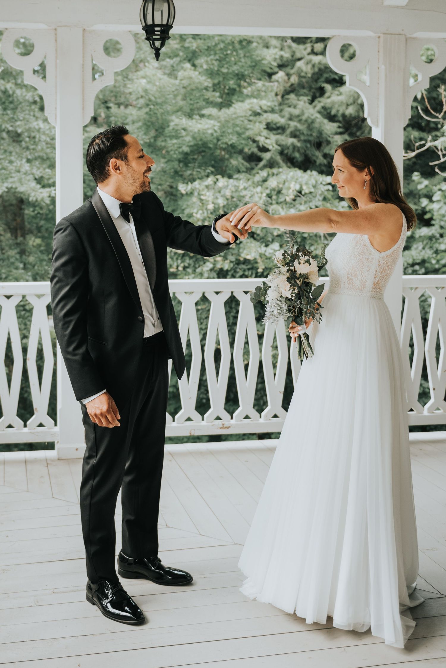 A bride in a flowing white gown and groom in tuxedo share a joyful moment on an elegant white porch during their wedding.