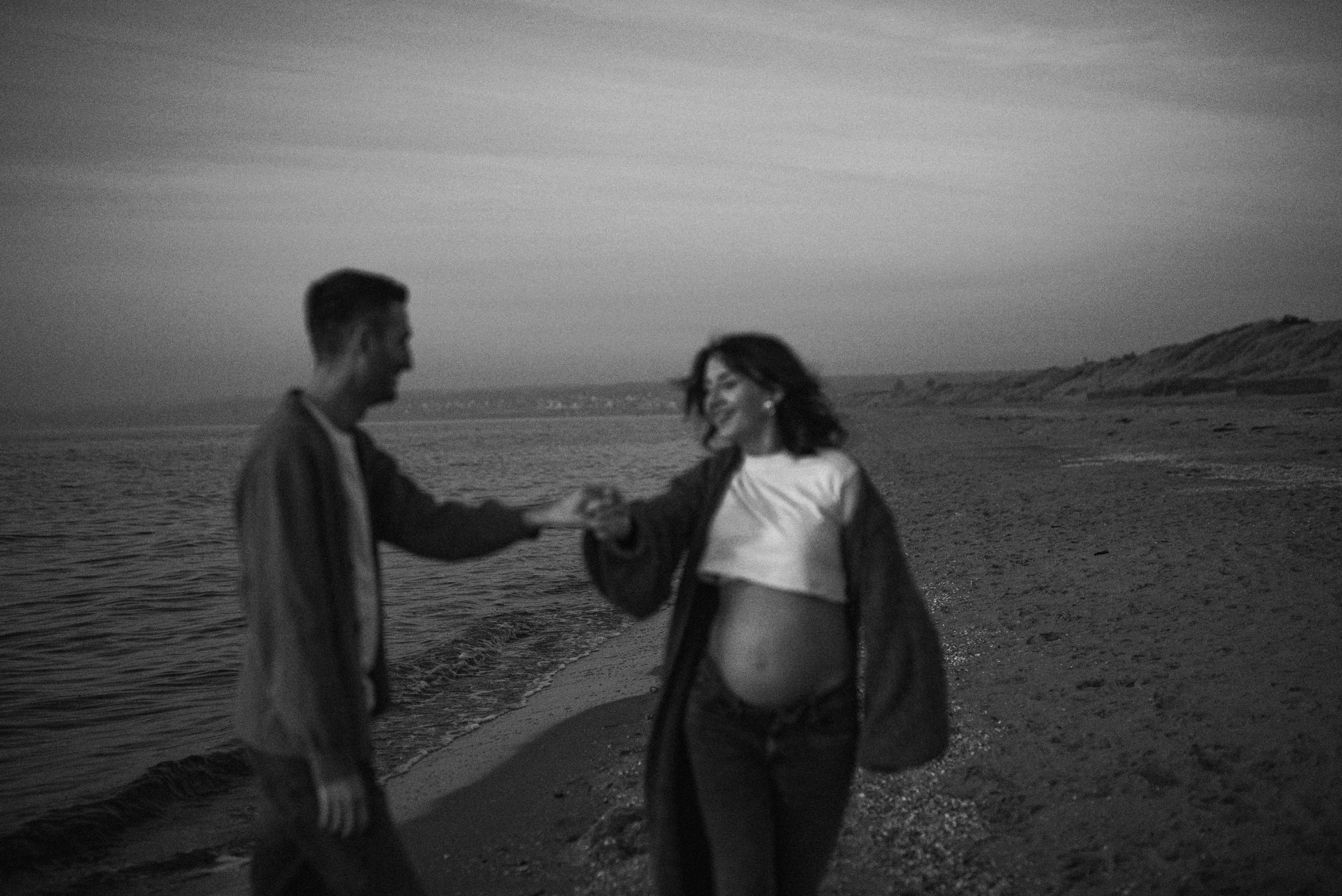 A black and white photo of a couple walking along a beach at sunset with mountains in the distance.