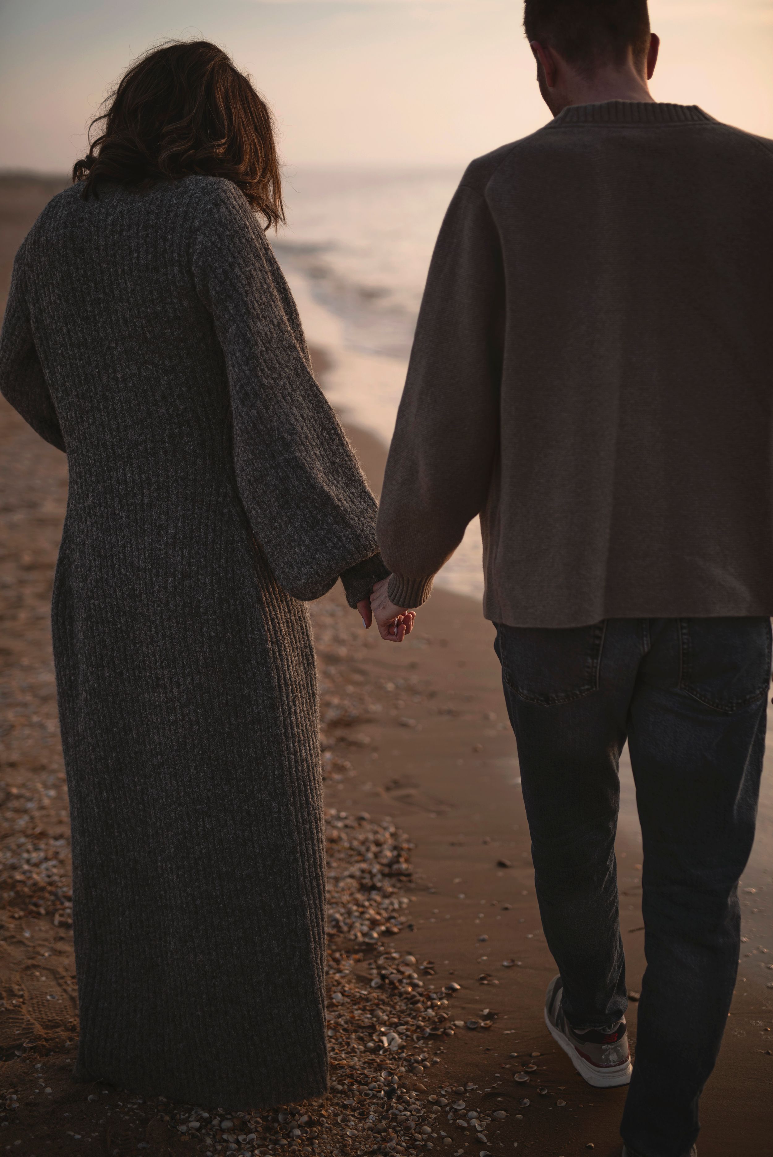 Two people hold hands while walking along a beach at sunset wearing cozy sweaters and casual attire.