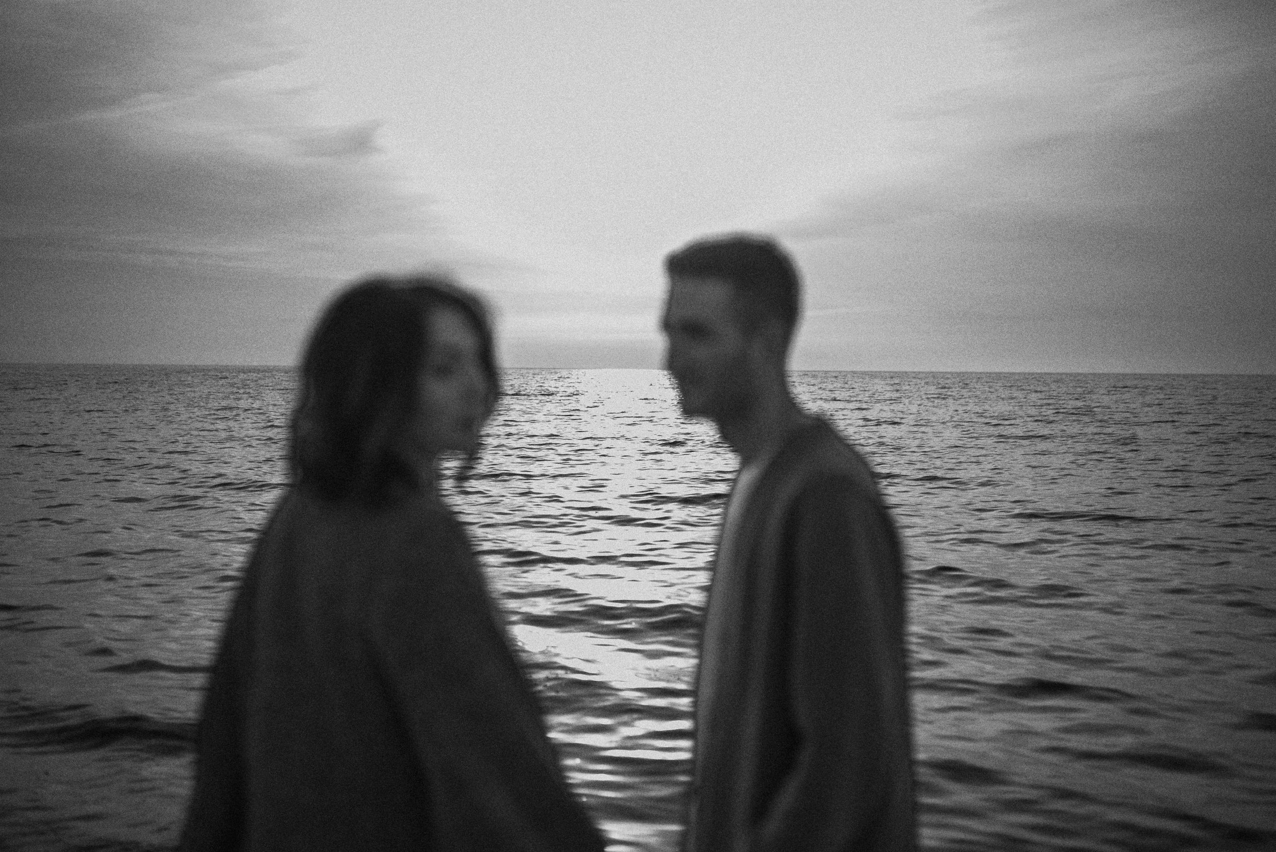 Black and white silhouette of a couple in profile standing near the ocean at sunset.