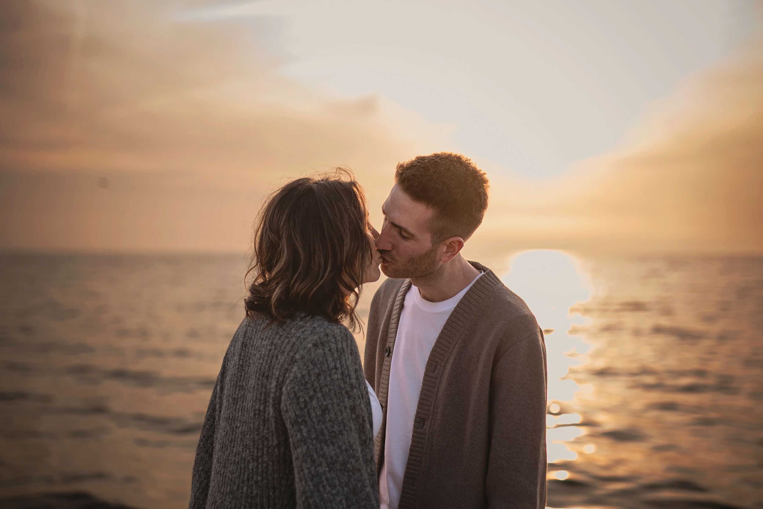 A couple shares a romantic moment at sunset by the ocean with golden light illuminating their silhouettes.
