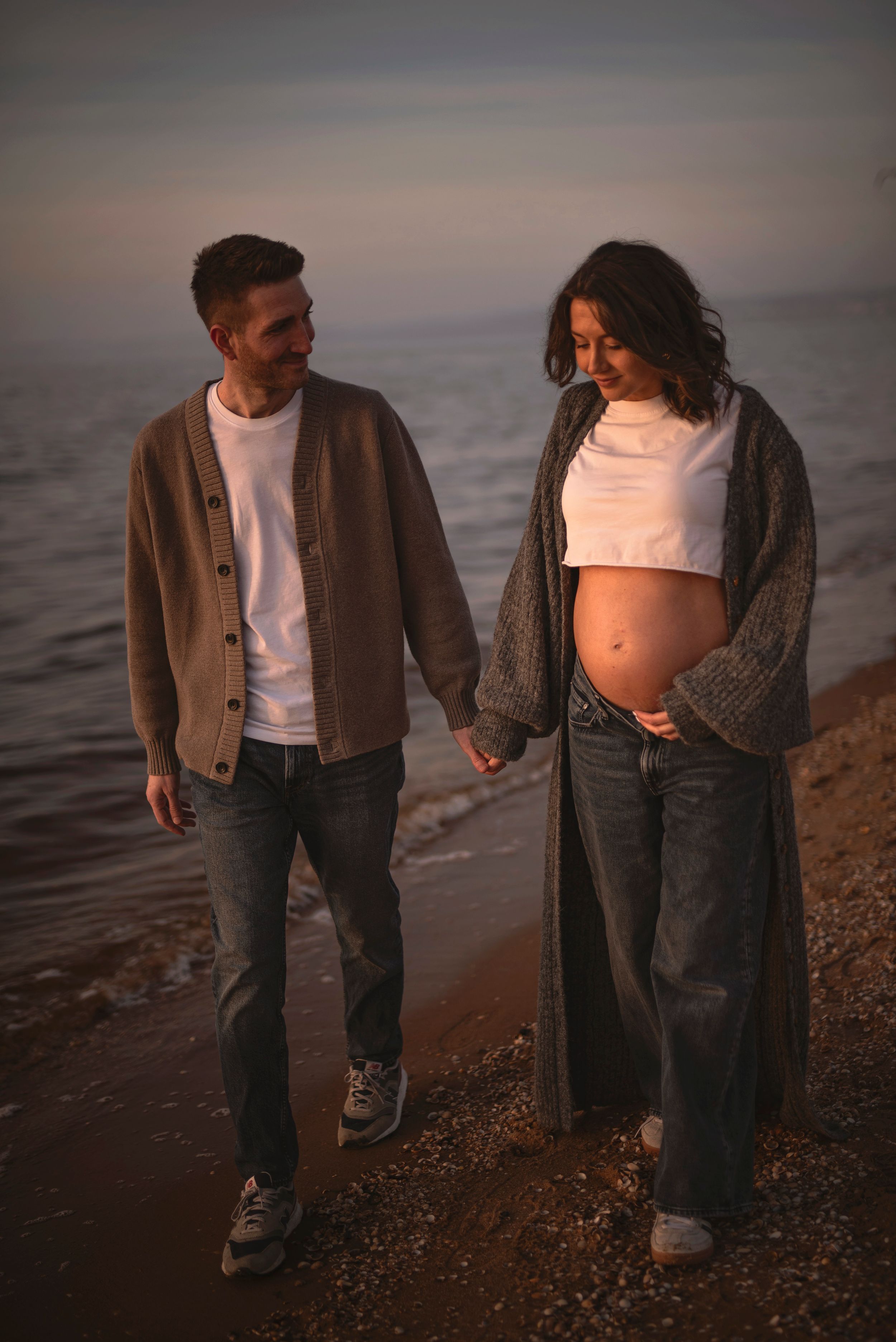 A couple strolls together on a beach at sunset with the woman showing off her pregnant belly in casual attire.