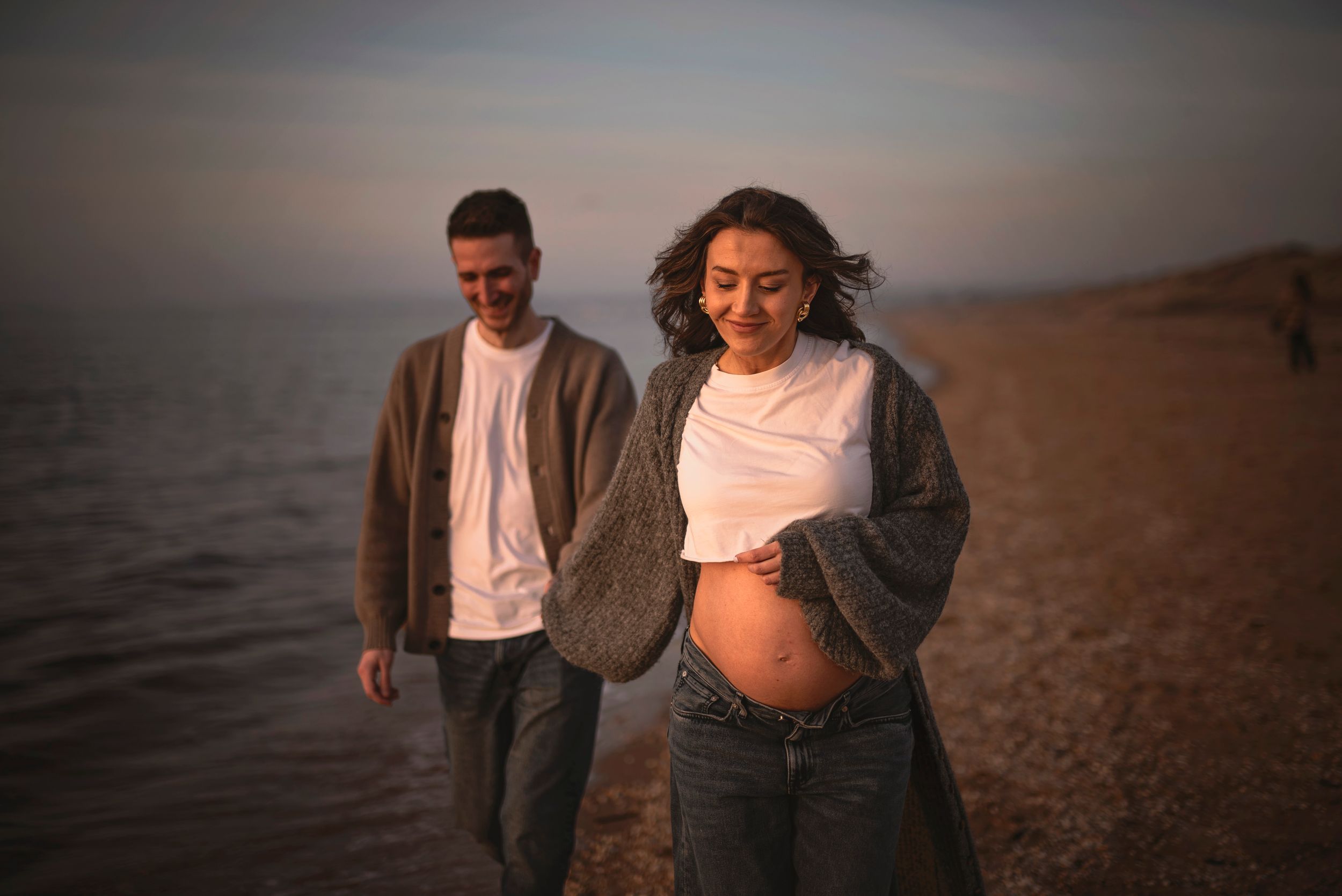 A romantic maternity photoshoot on a beach at sunset showing an expecting couple walking along the shore.