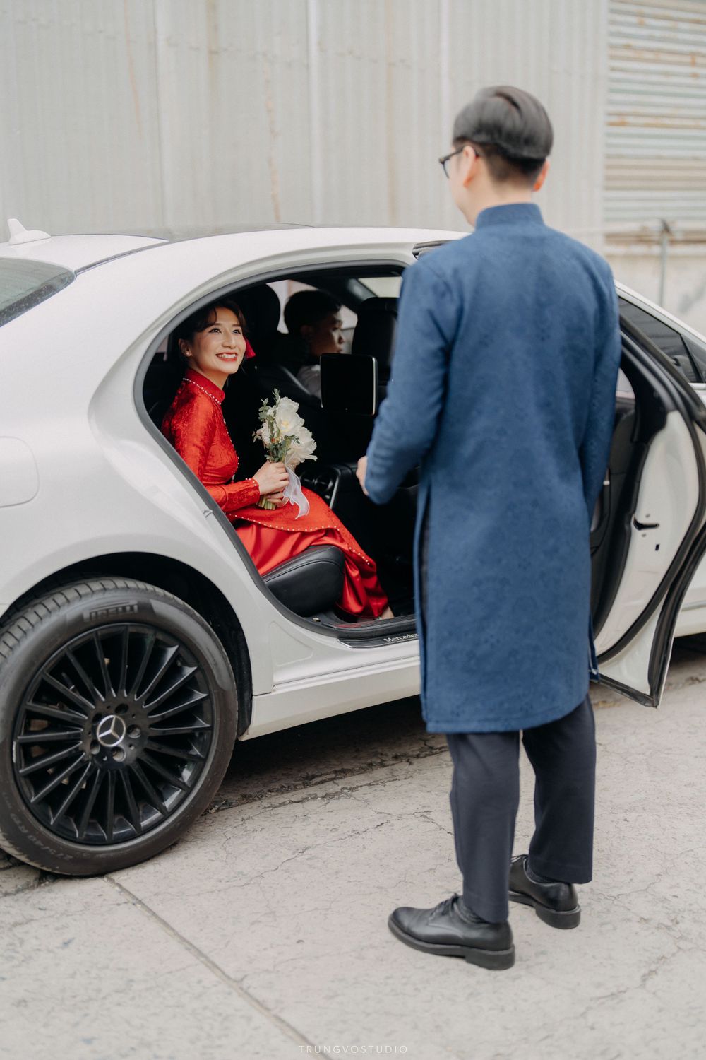 Vietnamese bride in red ao dai exits white Mercedes as groom in blue holds the door open.