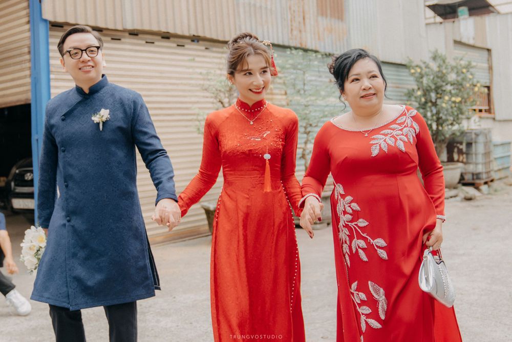 Vietnamese groom in blue ao dai walks with bride and mother in red ao dai at traditional wedding.