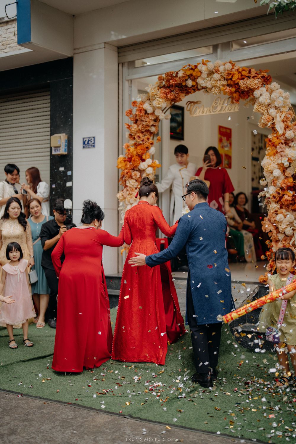 Vietnamese wedding couple in red and blue ao dai enter flower-adorned doorway with confetti celebration.