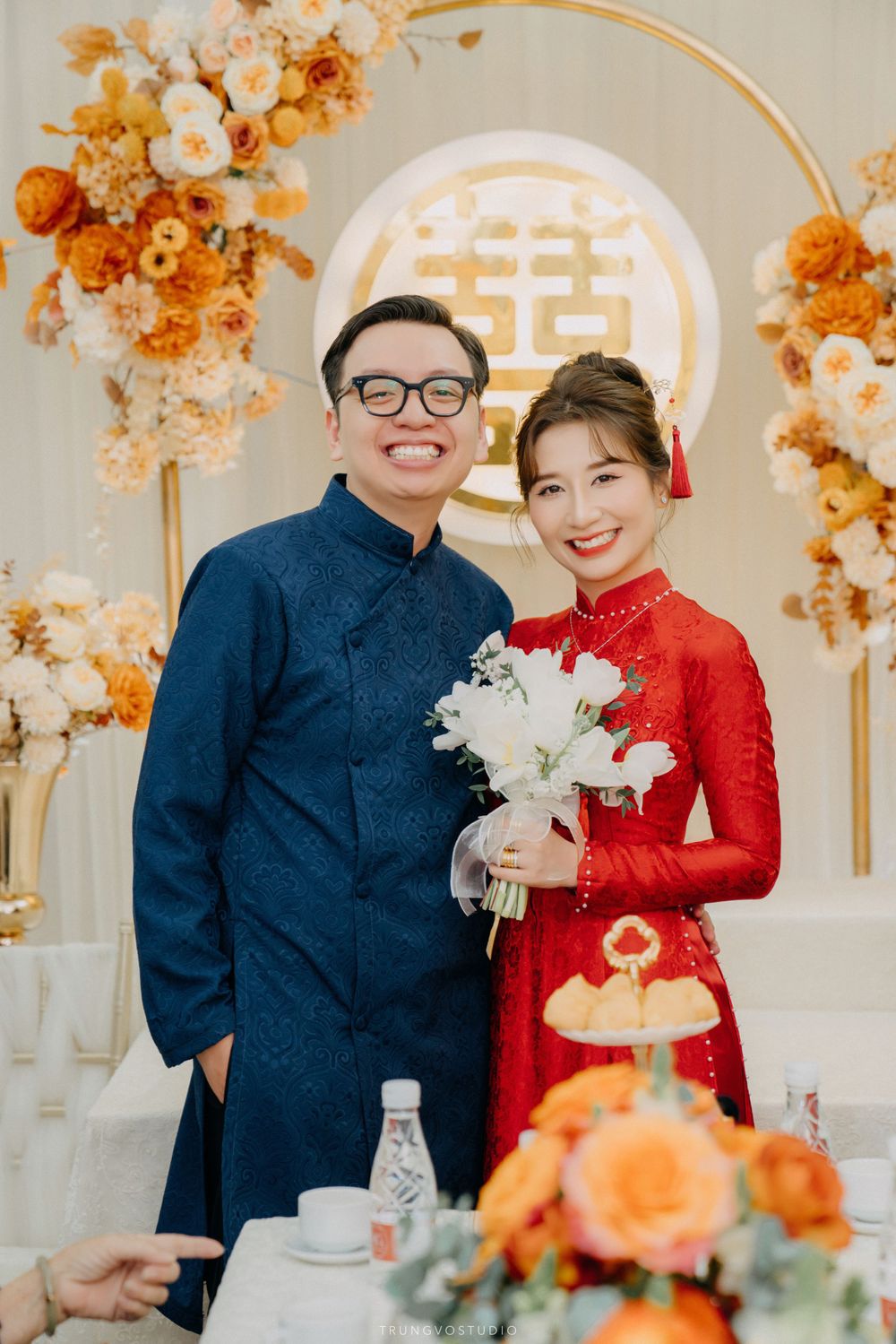 Vietnamese couple in traditional red and blue ao dai pose before golden double happiness wedding backdrop.