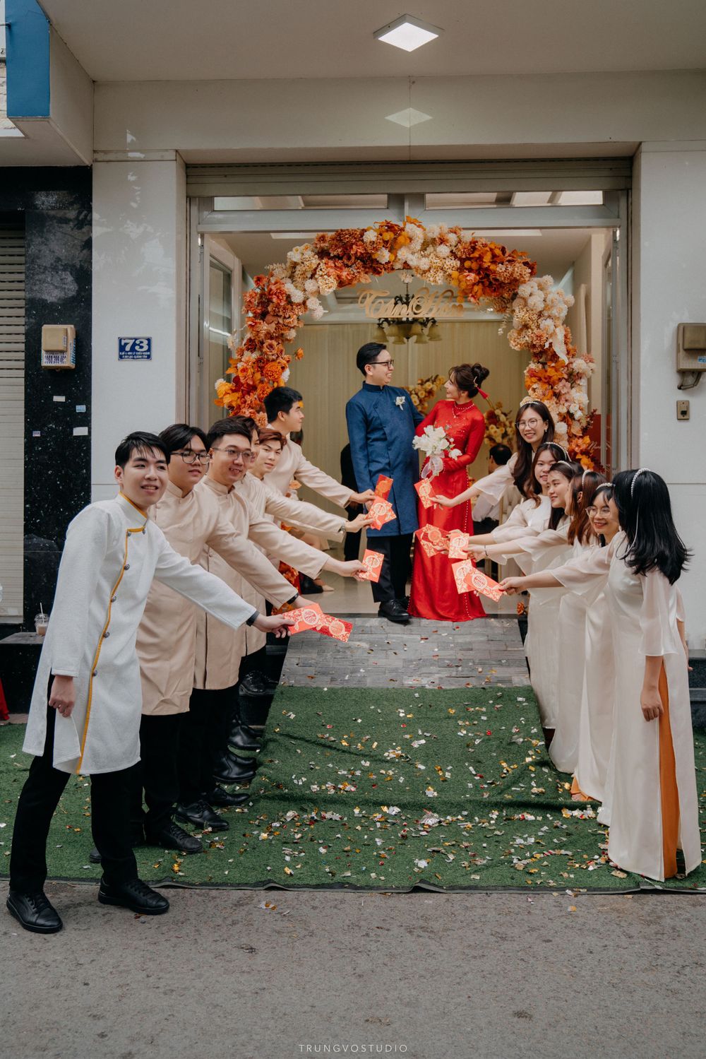 Vietnamese bridesmaids and groomsmen form a celebratory aisle for couple exiting flower-decorated doorway.
