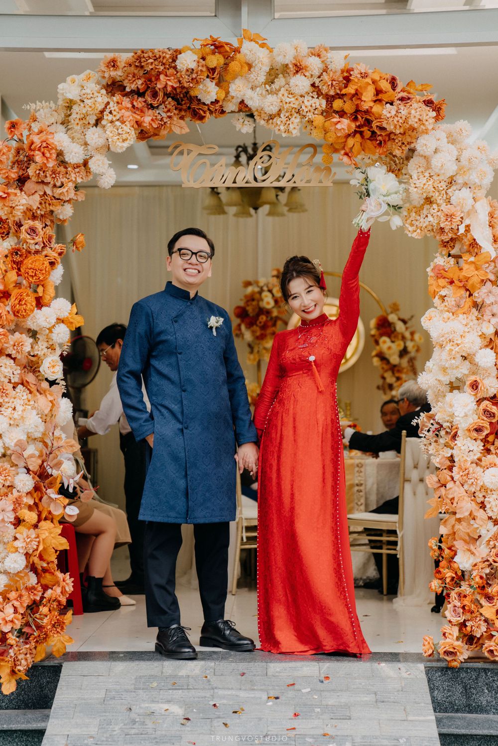 Vietnamese couple in traditional ao dai wedding attire pose under ornate orange and white floral arch.