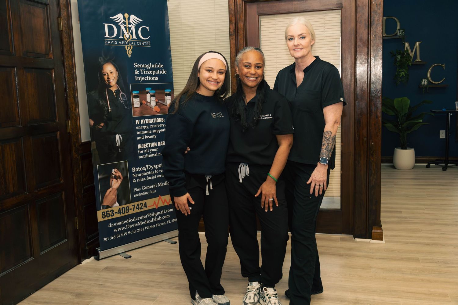 Three women in black scrubs smile together in front of a DMC wellness center banner in a professional office.