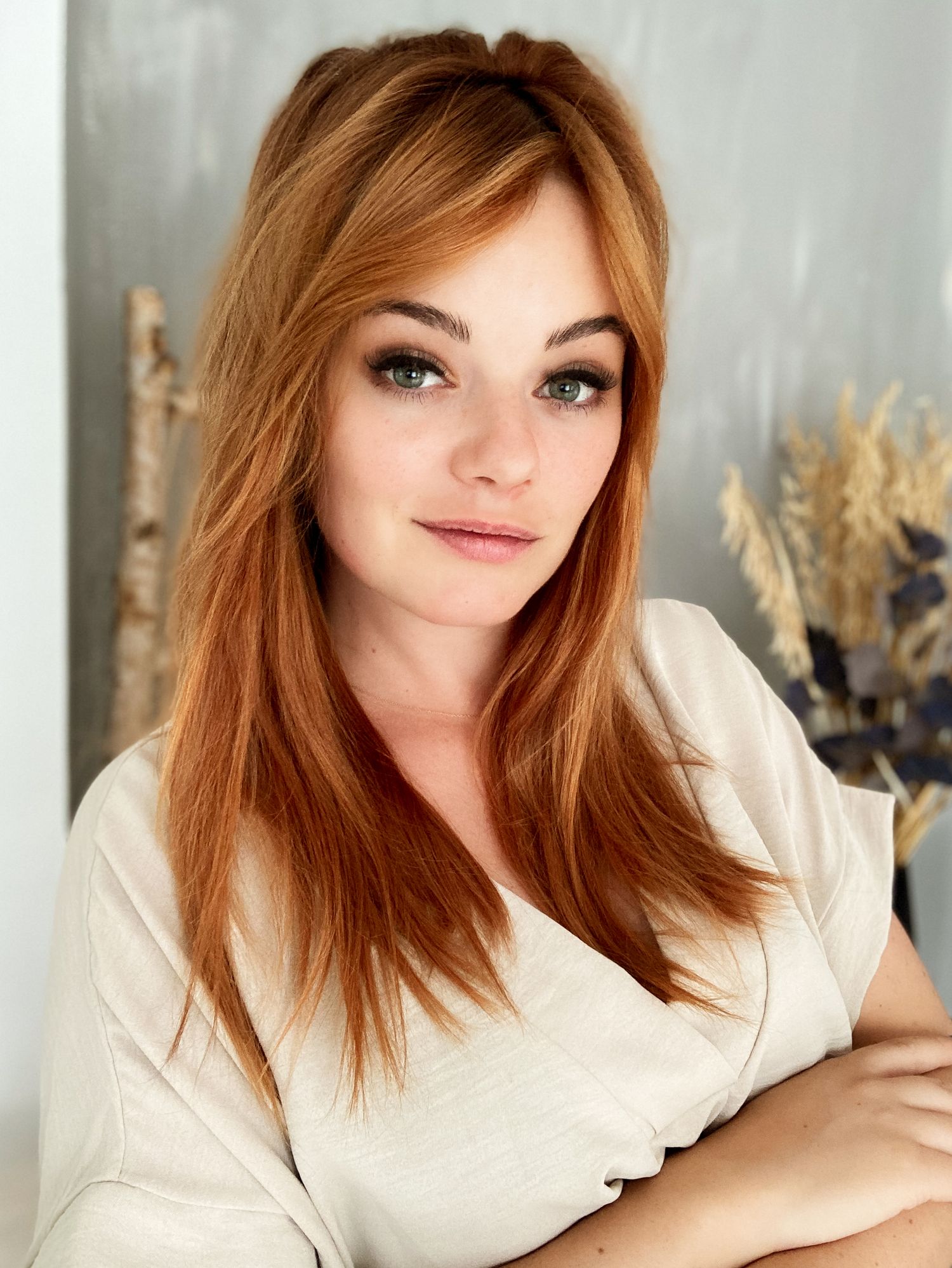 Portrait of someone with striking red hair wearing a cream colored top against a gray backdrop with dried flowers.