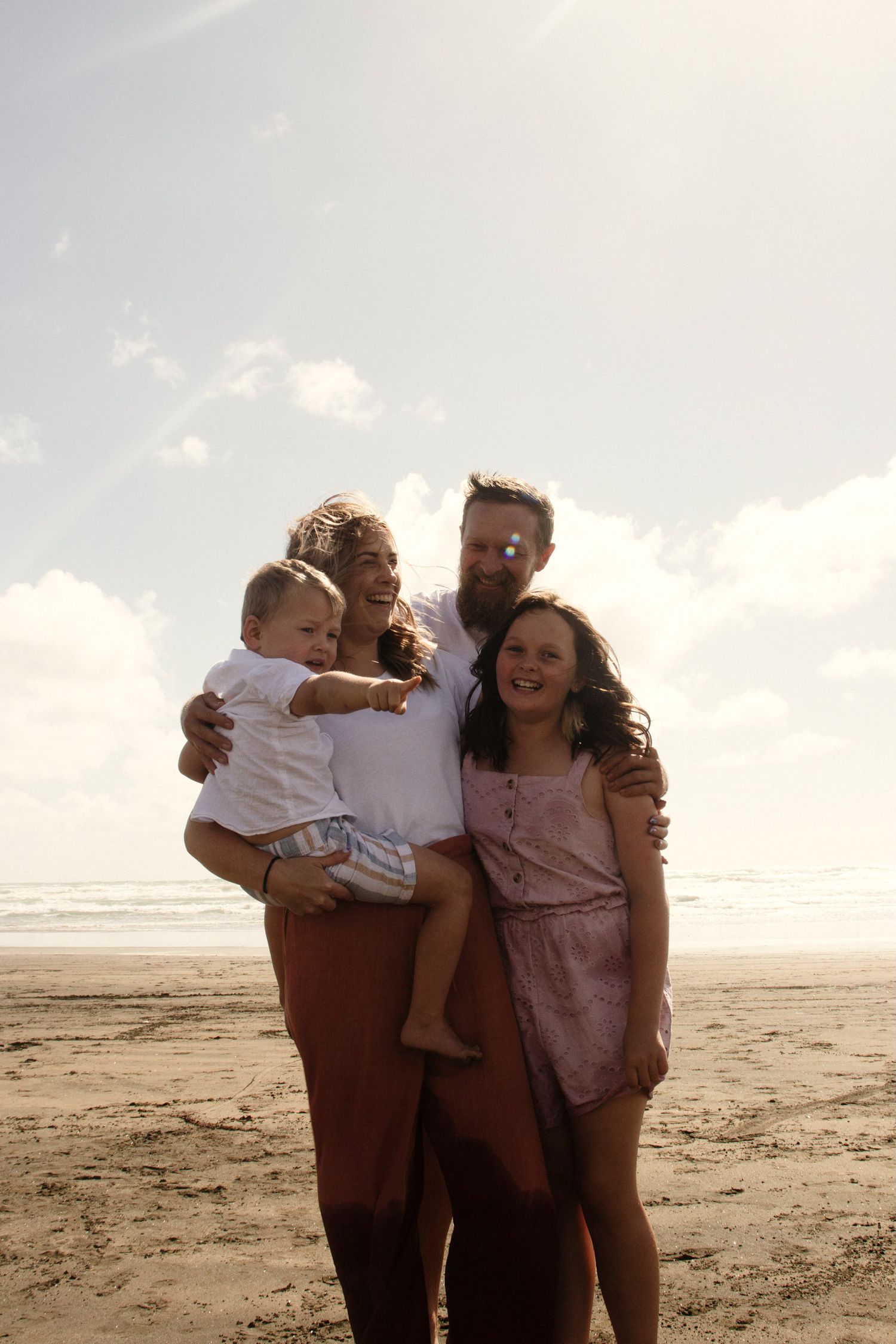 A family of four, mum, dad, little brother and big sister, at the beach at golden hour. Dad has has his arms wrapped around his family